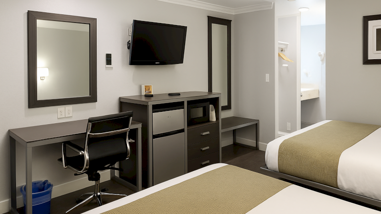 A modern hotel room with two single beds, a desk and chair, a wall-mounted TV, a mini-fridge and drawers, and a small bathroom doorway.