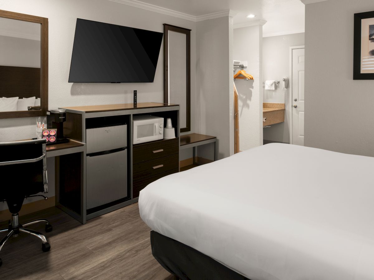 A modern hotel room with a desk, chair, flat-screen TV, mini-fridge, and a neatly made bed in muted tones.