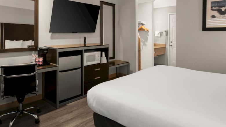 A modern hotel room with a desk, chair, flat-screen TV, mini-fridge, and a neatly made bed in muted tones.