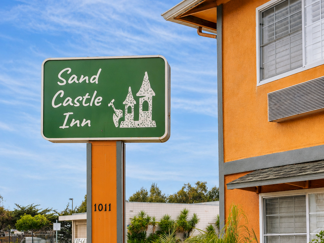 A roadside hotel sign for "Sand Castle Inn" with a green plaque, orange building, small trees, and clear blue sky above. End.
