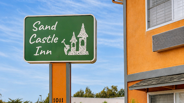 A roadside hotel sign for "Sand Castle Inn" with a green plaque, orange building, small trees, and clear blue sky above. End.