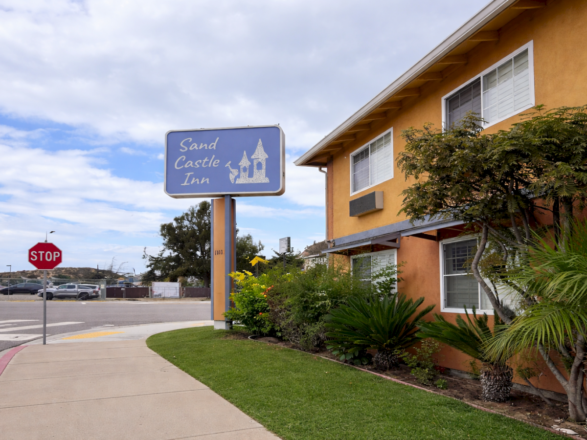 A roadside motel with a blue sign reading &ldquo;Sand Castle Inn,&rdquo; orange stucco building, palm trees, a manicured lawn, and a stop sign by the sidewalk.