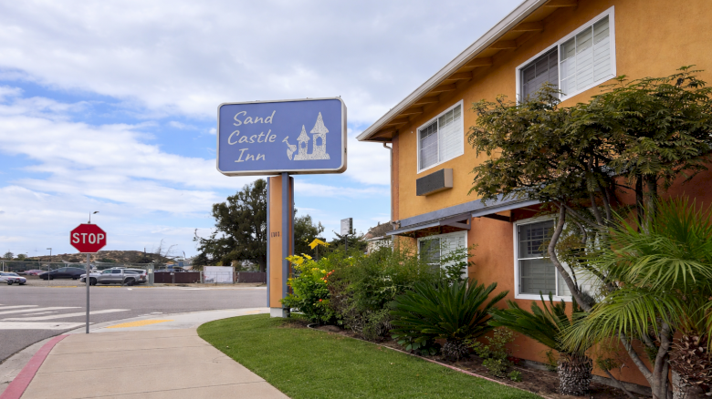 A roadside motel with a blue sign reading &ldquo;Sand Castle Inn,&rdquo; orange stucco building, palm trees, a manicured lawn, and a stop sign by the sidewalk.
